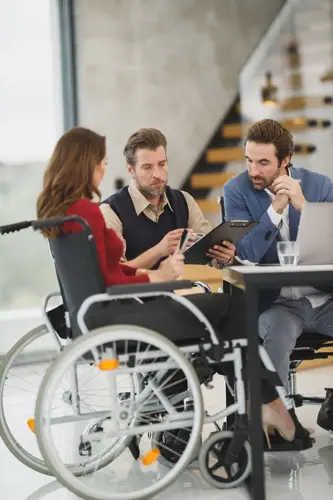 3 people, 2 men and a woman. The woman is in a wheelchair. they are at work by a table in conversation